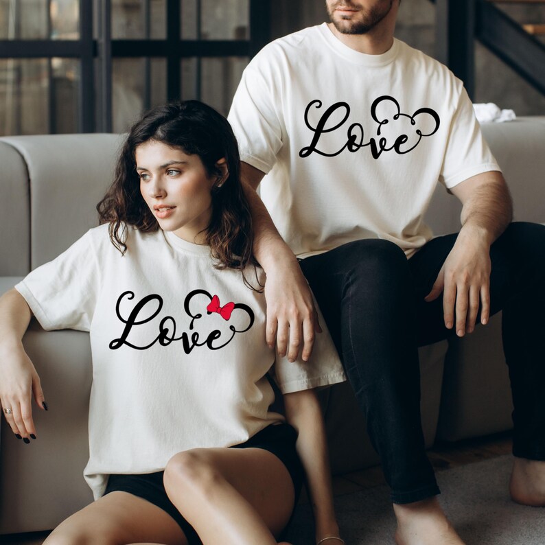 K&ouml;nnte beinhalten: Ein Paar tr&auml;gt wei&szlig;e T-Shirts mit dem Wort "Love" und einer Mickey-Maus-Silhouette. Das Shirt der Frau hat eine rote Schleife am Mickey-Maus-Ohr.