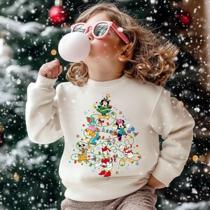 K&ouml;nnte beinhalten: Ein cremefarbenes Sweatshirt mit einem Weihnachtsbaum-Design aus Disney-Figuren. Der Baum enth&auml;lt Micky Maus, Minnie Maus, Donald Duck und Goofy. Das Kind auf dem Bild tr&auml;gt eine rosa Sonnenbrille und bl&auml;st eine Blase.