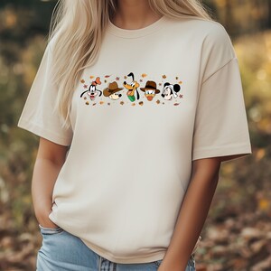 May include: Beige t-shirt featuring a cartoon graphic of Disney characters, including Goofy, Mickey Mouse, and Donald Duck, wearing hats and surrounded by autumn leaves. The shirt is a neutral color and has short sleeves.