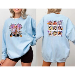 May include: Light blue sweatshirt featuring a Halloween-themed graphic on the front with a large bow, bats, and cartoon characters. The back of the sweatshirt displays a grid of cartoon characters in witch costumes. A casual, long-sleeved top.