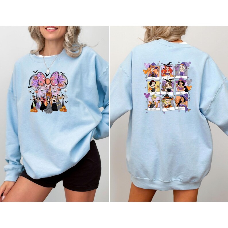 May include: Light blue sweatshirt featuring a Halloween-themed graphic on the front with a large bow, bats, and cartoon characters. The back of the sweatshirt displays a grid of cartoon characters in witch costumes. A casual, long-sleeved top.