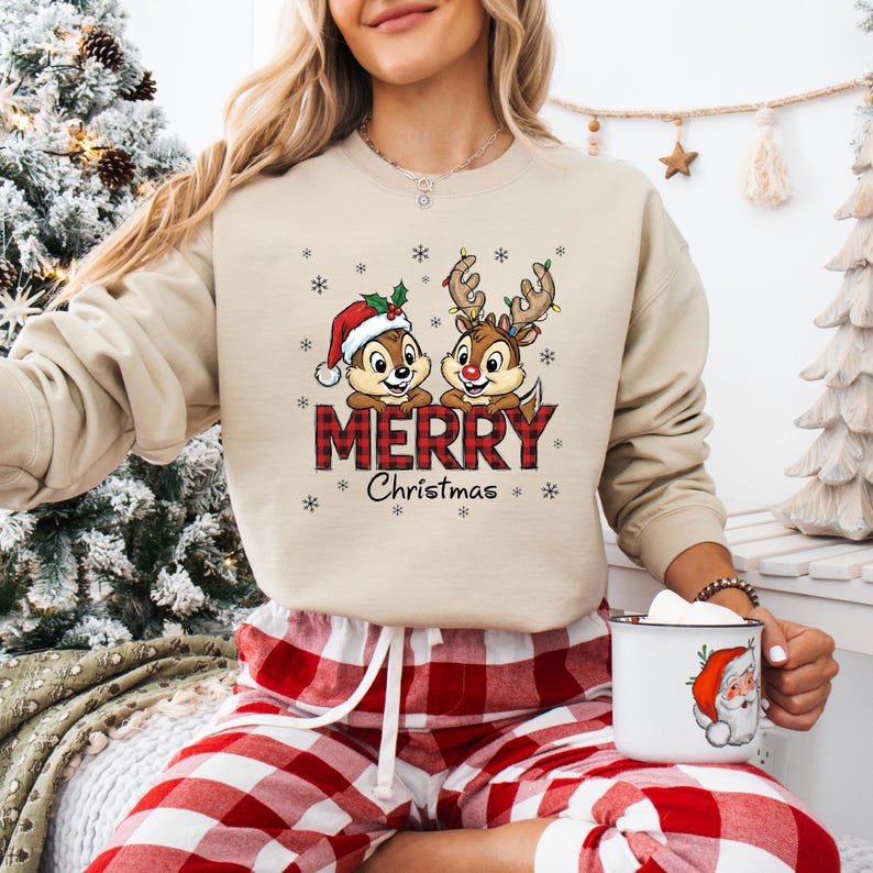 May include: A beige sweatshirt with a Christmas design. It features two cartoon chipmunks, one wearing a Santa hat and the other with reindeer antlers. The text "MERRY Christmas" is in a red plaid pattern, with snowflakes.