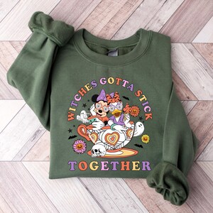 K&ouml;nnte beinhalten: Gr&uuml;nes Sweatshirt mit einem Aufdruck von Mickey Mouse und Daisy Duck, die als Hexen in einer Teetasse verkleidet sind. Der Text "Witches Gotta Stick Together" ist auf dem Sweatshirt aufgedruckt.