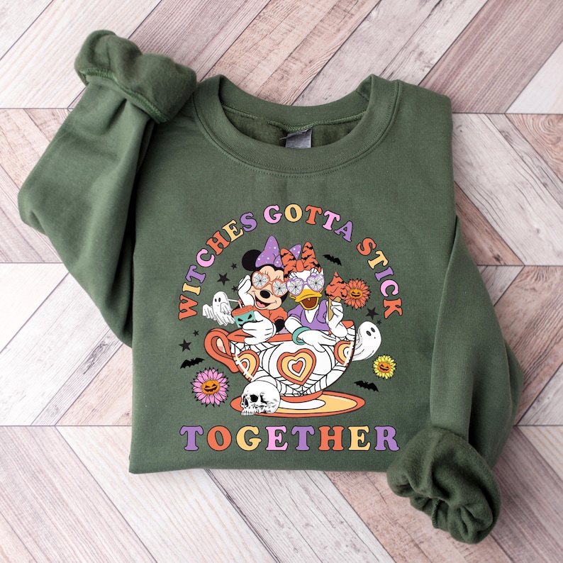 K&ouml;nnte beinhalten: Gr&uuml;nes Sweatshirt mit einem Aufdruck von Mickey Mouse und Daisy Duck, die als Hexen in einer Teetasse verkleidet sind. Der Text "Witches Gotta Stick Together" ist auf dem Sweatshirt aufgedruckt.