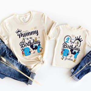 May include: Two cream colored t-shirts with a Mickey Mouse theme. The larger shirt says "Mommy of the Birthday Boy" with Mickey Mouse and friends. The smaller shirt says "2 Birthday Boy" with Mickey Mouse and friends and the name "Jordan".