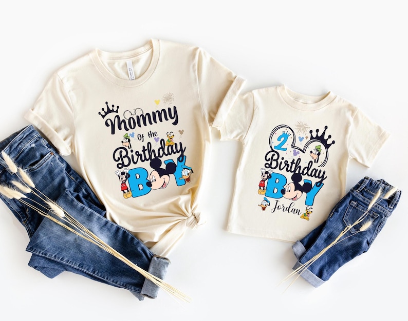May include: Two cream colored t-shirts with a Mickey Mouse theme. The larger shirt says "Mommy of the Birthday Boy" with Mickey Mouse and friends. The smaller shirt says "2 Birthday Boy" with Mickey Mouse and friends and the name "Jordan".