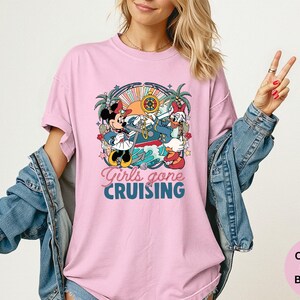 May include: A light pink t-shirt featuring a graphic of Mickey and Minnie Mouse, and Donald Duck on a boat, with the text "Girls gone CRUISING". The shirt is part of the Comfort Colors Blossom collection.