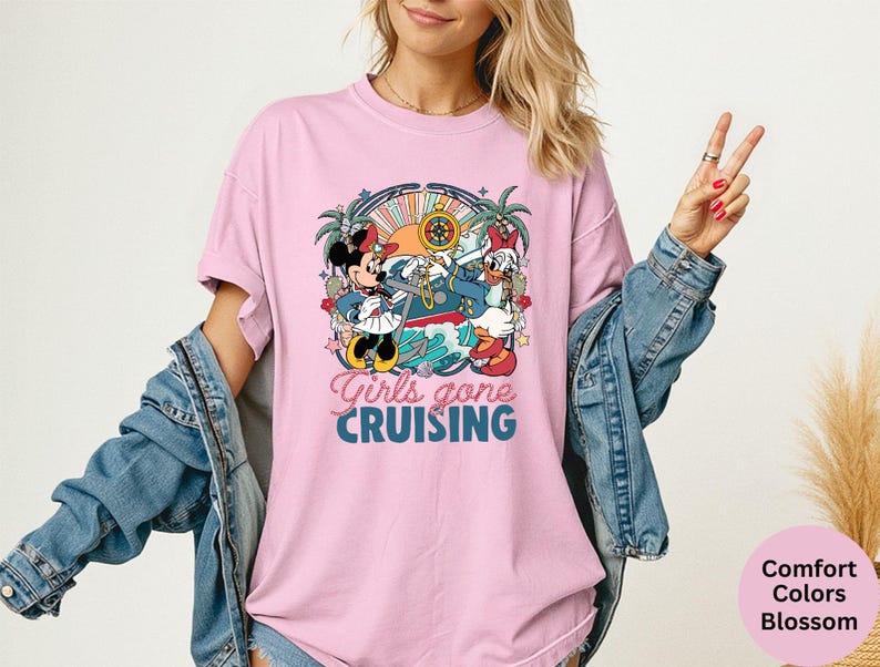 May include: A light pink t-shirt featuring a graphic of Mickey and Minnie Mouse, and Donald Duck on a boat, with the text "Girls gone CRUISING". The shirt is part of the Comfort Colors Blossom collection.