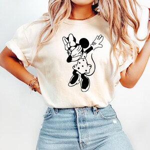 May include: A white t-shirt with a black and white illustration of Minnie Mouse doing the dab. Minnie Mouse is wearing a polka dot dress and a bow.