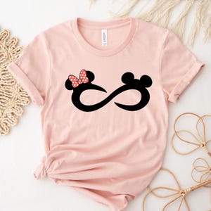 May include: A pink t-shirt featuring a black infinity symbol and a cartoon image of Mickey Mouse and Minnie Mouse. Minnie Mouse is wearing a red bow with white polka dots.
