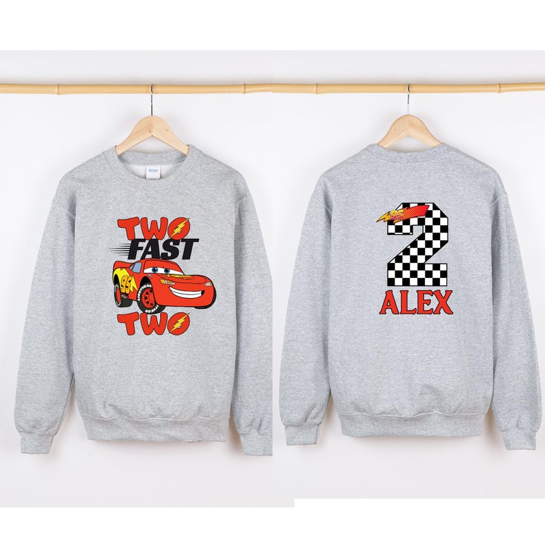 K&ouml;nnte beinhalten: Zwei graue Sweatshirts mit unterschiedlichen Designs. Eines zeigt ein rotes Cartoon-Auto und den Text "TWO FAST TWO". Das andere hat eine karierte Zahl 2 und den Namen "ALEX" in Rot.
