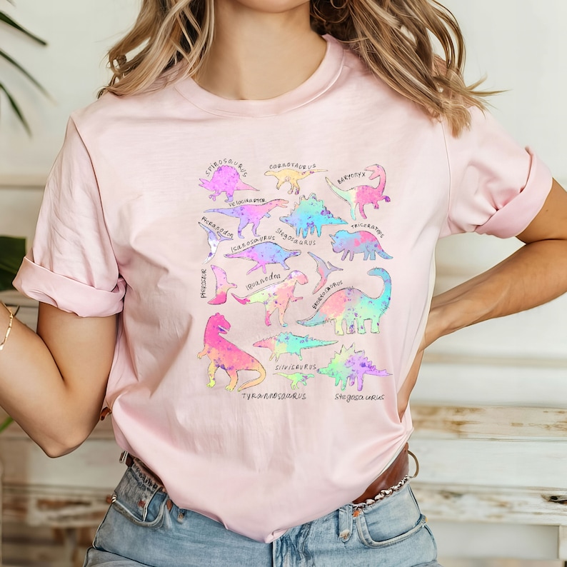 May include: A pink t-shirt with a rainbow design featuring various dinosaurs with their names printed below each one. The dinosaurs include Triceratops, Stegosaurus, Tyrannosaurus Rex, Brachiosaurus, and more.