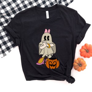 Puede incluir: Camiseta negra con un fantasma de dibujos animados que lleva un lazo rosa y sostiene un cubo de calabaza. El fantasma tiene una s&aacute;bana blanca y sostiene sus brazos frente a &eacute;l.