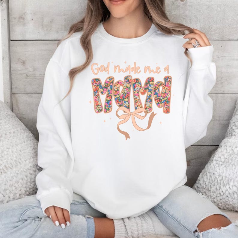 Pu&ograve; includere: Felpa bianca con la scritta "God made me a Mama" in un design floreale. La parola "Mama" &egrave; riempita di fiori colorati. Un fiocco color pesca &egrave; sotto il testo. La felpa &egrave; in stile casual.