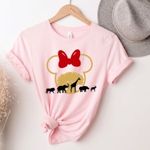 May include: Pink t-shirt with a gold Mickey Mouse head silhouette. Inside the ears is a gold tree with a giraffe, elephant, lion, hippopotamus, and a goat. The bow on top of Mickey's head is red.