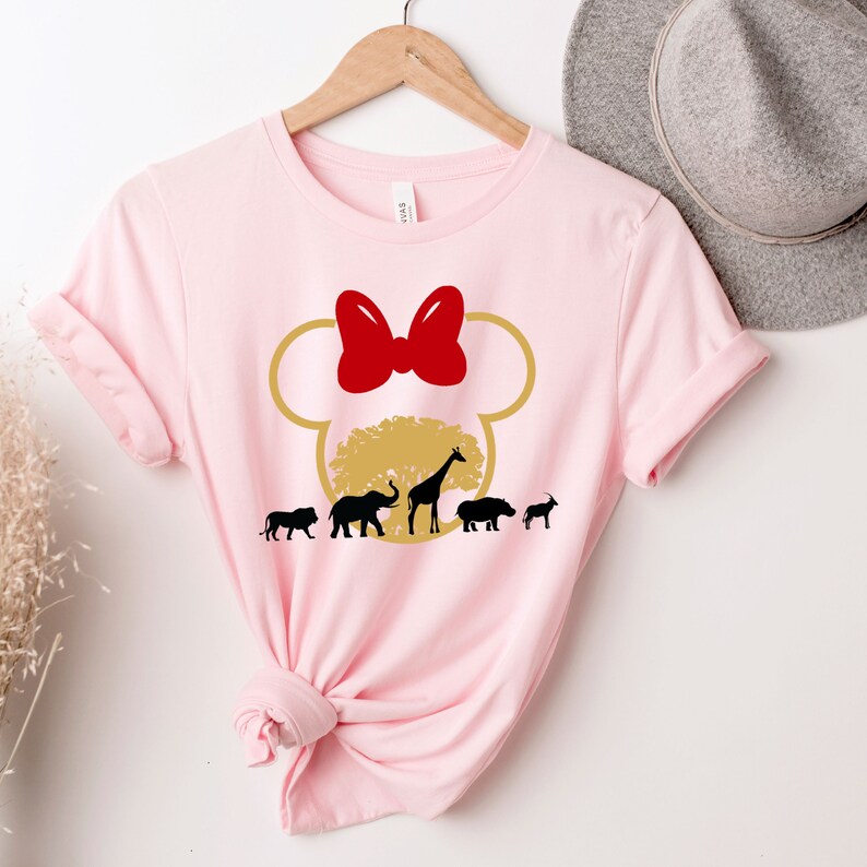 May include: Pink t-shirt with a gold Mickey Mouse head silhouette. Inside the ears is a gold tree with a giraffe, elephant, lion, hippopotamus, and a goat. The bow on top of Mickey's head is red.