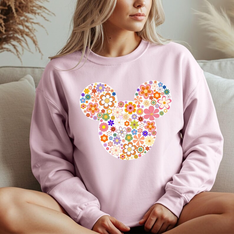 May include: A light pink sweatshirt featuring a floral Mickey Mouse design. The design is composed of various colorful flowers in shades of pink, orange, yellow, blue, and green. The sweatshirt has a crew neck and long sleeves.