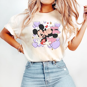May include: A white t-shirt with a graphic print of Mickey Mouse and Minnie Mouse kissing. The graphic is surrounded by purple hearts and gold glitter.