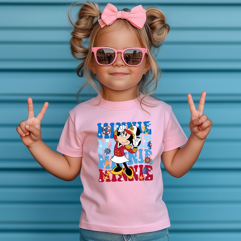 May include: A pink t-shirt with a graphic of Minnie Mouse dressed as a sailor. The graphic includes the text "Minnie Mouse Minnie" in a retro font. The shirt is being worn by a child with pink sunglasses and a pink bow in their hair.