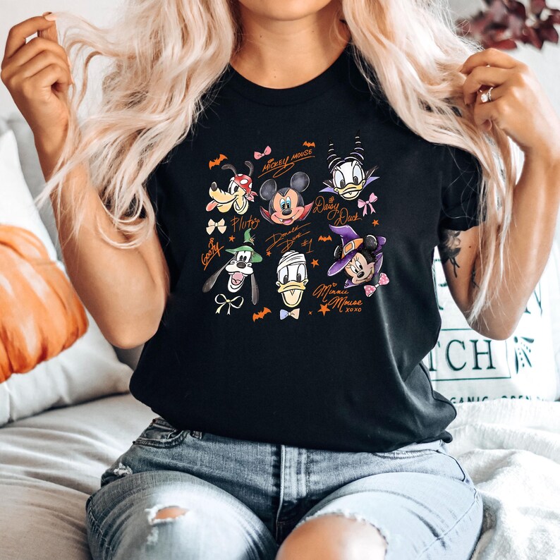 Puede incluir: Camiseta negra con un dise&ntilde;o de Halloween con personajes de dibujos animados como Mickey Mouse, Minnie Mouse, Donald Duck y Goofy. El dise&ntilde;o incluye murci&eacute;lagos, lazos y los nombres de los personajes.