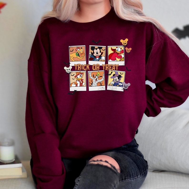 Op de afbeelding: Een bordeauxrood sweatshirt met een grafische print van Mickey Mouse en andere Disney-figuren in een Halloween-thema. De tekst "Trick or Treat" is op het sweatshirt gedrukt.