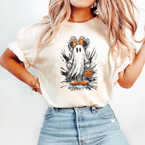 Peut inclure: T-shirt cr&egrave;me avec un motif d'Halloween. Le motif repr&eacute;sente un fant&ocirc;me avec des oreilles de souris et un n&oelig;ud, tenant un seau de citrouille. Le design est en noir et blanc avec des accents orange, sur un fond &eacute;clabouss&eacute;.