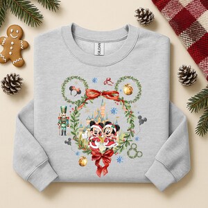 May include: A light gray sweatshirt featuring a festive Mickey and Minnie Mouse design. The design includes a wreath, castle, snowflakes, and a red bow. The sweatshirt has long sleeves and a crew neck.