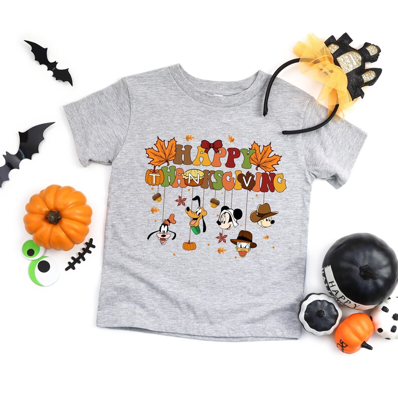 Op de afbeelding: Een lichtgrijs t-shirt met een kleurrijke "Happy Thanksgiving" afbeelding met cartoonfiguren en herfstbladeren. Het shirt is omgeven door Halloween- en Thanksgiving-decoraties, waaronder pompoenen en vleermuizen.
