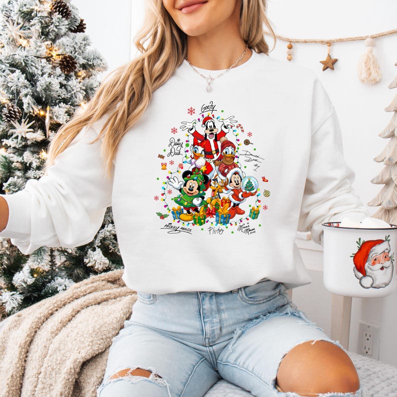 K&ouml;nnte beinhalten: Wei&szlig;es Sweatshirt mit einem festlichen Weihnachtsdesign mit Mickey Mouse, Donald Duck, Goofy und anderen Disney-Figuren. Die Grafik enth&auml;lt farbenfrohe Konfetti, Geschenke und die Unterschriften der Charaktere. Eine Santa-Tasse ist sichtbar.