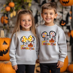 Puede incluir: Dos ni&ntilde;os con sudaderas grises claras con un gr&aacute;fico colorido. El gr&aacute;fico presenta orejas de Mickey Mouse, un castillo y la palabra "THANKFUL" en naranja y amarillo. La sudadera de la ni&ntilde;a tiene un lazo amarillo.