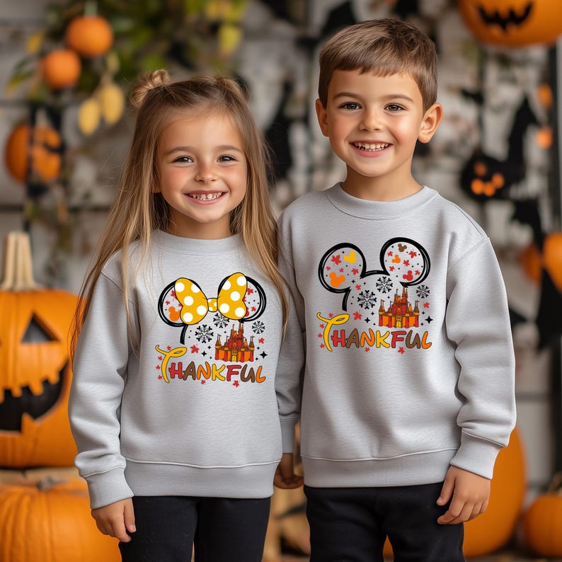 Puede incluir: Dos ni&ntilde;os con sudaderas grises claras con un gr&aacute;fico colorido. El gr&aacute;fico presenta orejas de Mickey Mouse, un castillo y la palabra "THANKFUL" en naranja y amarillo. La sudadera de la ni&ntilde;a tiene un lazo amarillo.