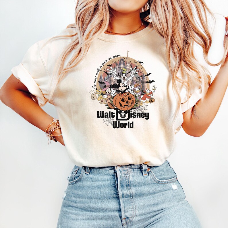 Puede incluir: Camiseta color crema con un dise&ntilde;o tem&aacute;tico de Halloween con personajes de Disney, una calabaza tallada y un castillo. El texto "Walt Disney World" se muestra debajo del dise&ntilde;o, con la frase "The most magical place on earth" arriba.