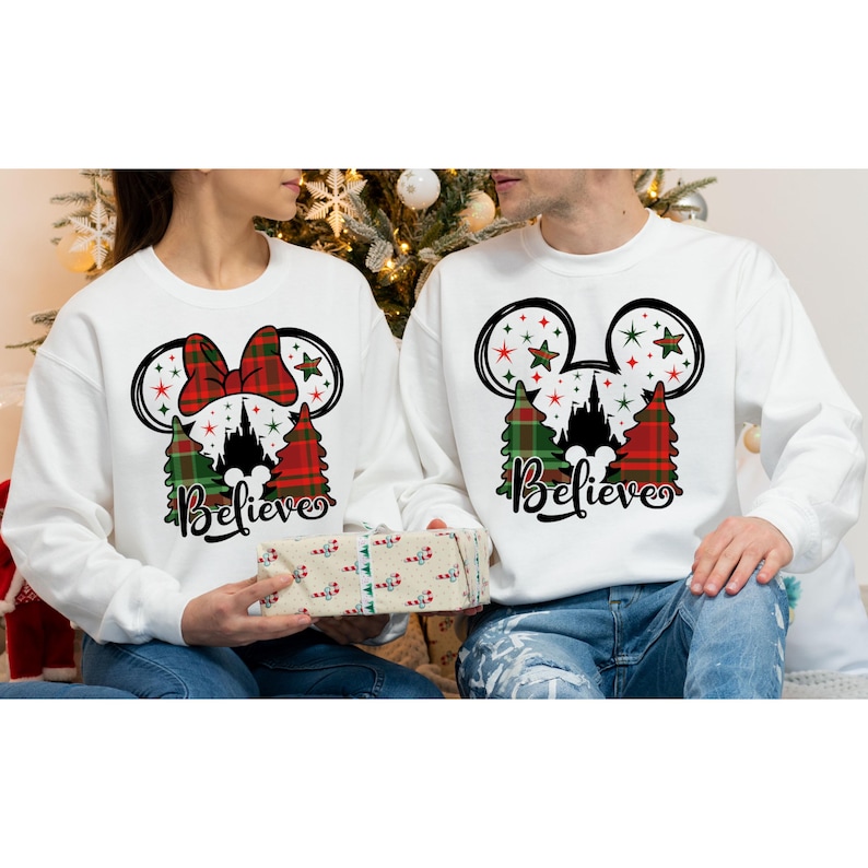 May include: Two white sweatshirts with a Christmas theme. The sweatshirts feature a Mickey Mouse head with a red bow and green and red plaid trees. The text 'Believe' is printed below the image. The sweatshirts are worn by two people.