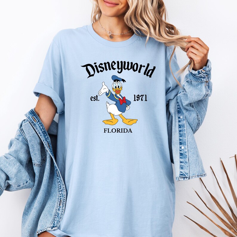 May include: Light blue t-shirt featuring the text "Disneyworld" above a cartoon image of Donald Duck. The text "est. 1971 FLORIDA" is below the image. The shirt has rolled-up sleeves.