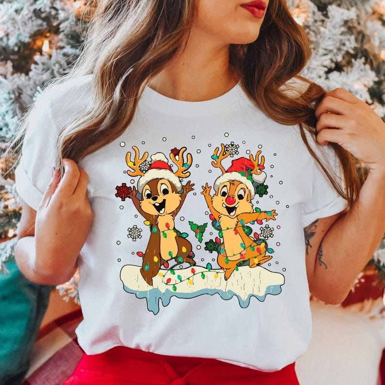 May include: White t-shirt featuring a Christmas design with two cartoon chipmunks wearing Santa hats and reindeer antlers. The chipmunks are decorated with Christmas lights and are standing on a snowy surface.