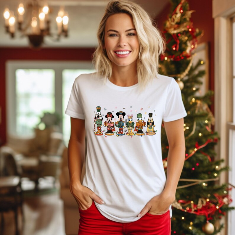 May include: White t-shirt featuring a line of cartoon nutcracker characters. The characters are in various colors and designs, including Mickey Mouse and Donald Duck. The shirt is a crew neck style.