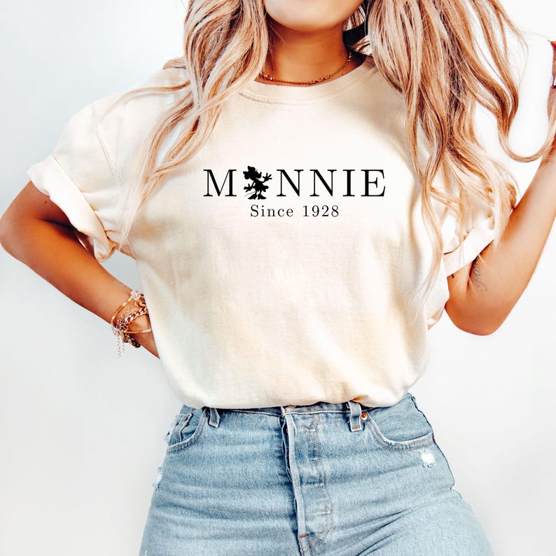 May include: Cream-colored t-shirt with the word "Minnie" in black, featuring a silhouette of a character and the text "Since 1928." The shirt is paired with blue denim shorts. The overall style is casual and comfortable.