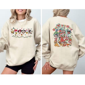 Op de afbeelding: Beige sweatshirt met een feestelijk Disney-ontwerp. De voorkant toont Mickey Mouse, Minnie Mouse, Donald Duck en Goofy in vakantiekleding. De achterkant toont een kleurrijke illustratie van Disney-personages en de tekst "Walt Disney World".