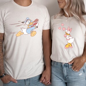 May include: A matching pair of t-shirts featuring a cartoon image of Donald Duck running with a bouquet of flowers and Daisy Duck standing with hearts around her.