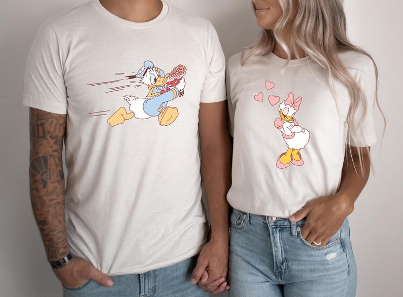 May include: A matching pair of t-shirts featuring a cartoon image of Donald Duck running with a bouquet of flowers and Daisy Duck standing with hearts around her.