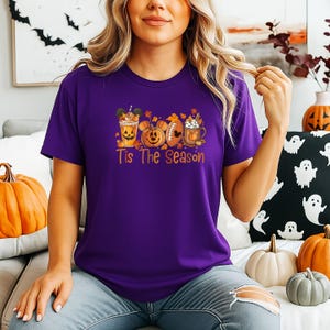 K&ouml;nnte beinhalten: Ein lila T-Shirt mit einem herbstlichen Motiv, das einen K&uuml;rbis, einen Football und eine Kaffeetasse zeigt. Der Text "Tis The Season" ist darunter gedruckt. Das Shirt ist aus einem weichen Material.