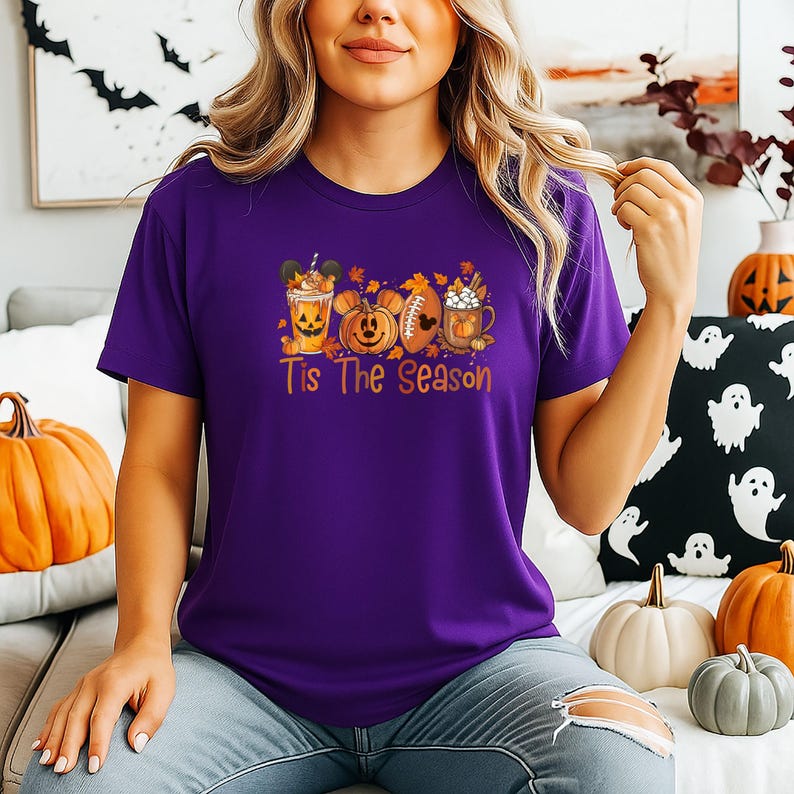K&ouml;nnte beinhalten: Ein lila T-Shirt mit einem herbstlichen Motiv, das einen K&uuml;rbis, einen Football und eine Kaffeetasse zeigt. Der Text "Tis The Season" ist darunter gedruckt. Das Shirt ist aus einem weichen Material.