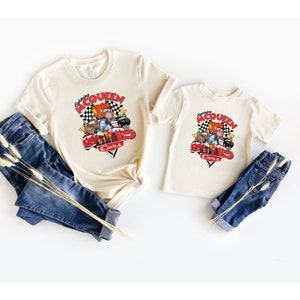 May include: Two beige t-shirts with a graphic of Lightning McQueen and other Cars characters. The text on the shirts reads "Racing McQueen Cars".