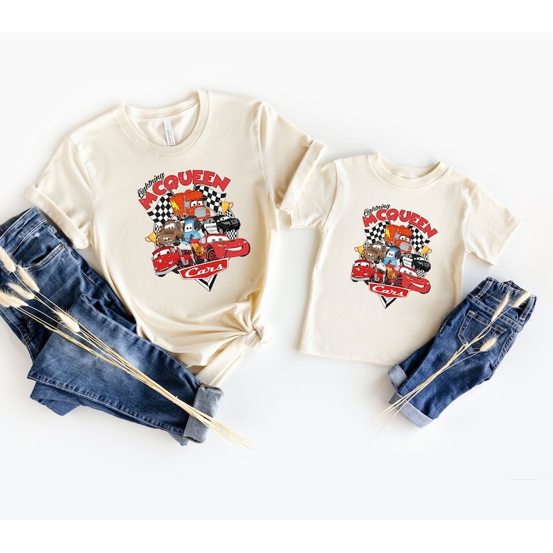 May include: Two beige t-shirts with a graphic of Lightning McQueen and other Cars characters. The text on the shirts reads "Racing McQueen Cars".
