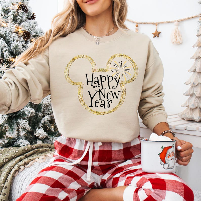 May include: A beige sweatshirt with a gold glitter Mickey Mouse silhouette and the text "Happy New Year" in black. The sweatshirt is worn by a person wearing red and white plaid pajama pants.