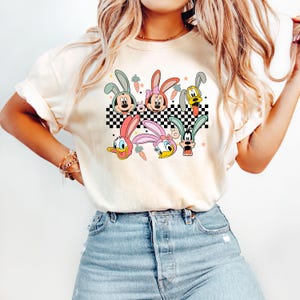 May include: A white t-shirt with a checkered pattern and cartoon characters wearing bunny ears. The characters include Mickey Mouse, Minnie Mouse, Donald Duck, Daisy Duck, and Goofy.