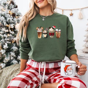 May include: An olive green sweatshirt featuring a graphic of five festive coffee drinks with Mickey Mouse ears and holiday accents. The person is wearing red and white plaid pajama pants and holding a white mug with a Santa Claus graphic.