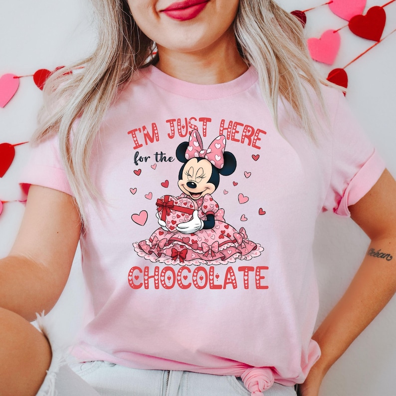 May include: Pink t-shirt featuring a graphic of Minnie Mouse holding a heart-shaped gift box. The text "I'M JUST HERE for the CHOCOLATE" is printed on the shirt. Hearts are scattered around the Minnie Mouse graphic.