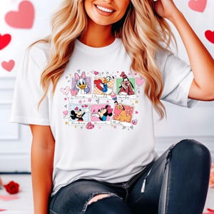 K&ouml;nnte beinhalten: Wei&szlig;es T-Shirt mit einem Grafikdruck von Mickey Mouse, Minnie Mouse, Pluto, Donald Duck, Daisy Duck und Goofy in einer Collage von Polaroid-Rahmen mit Herzen und Sternen. Der Text "Mickey", "Minnie", "Pluto", "Daisy", "Donald" und "Goofy" ist unter jedem Rahmen gedruckt.