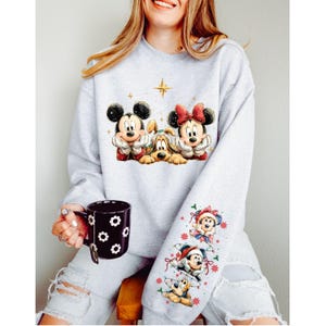 May include: A light gray sweatshirt featuring Mickey Mouse, Pluto, and Minnie Mouse in a Christmas design. The sleeve has additional Christmas-themed character graphics. A black mug with white flower designs is held in the hand.